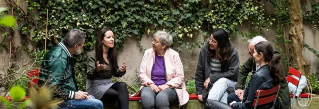Un groupe de six personnes de différents âges est assis en cercle dans un jardin verdoyant, écoutant attentivement une femme qui s'exprime avec animation.